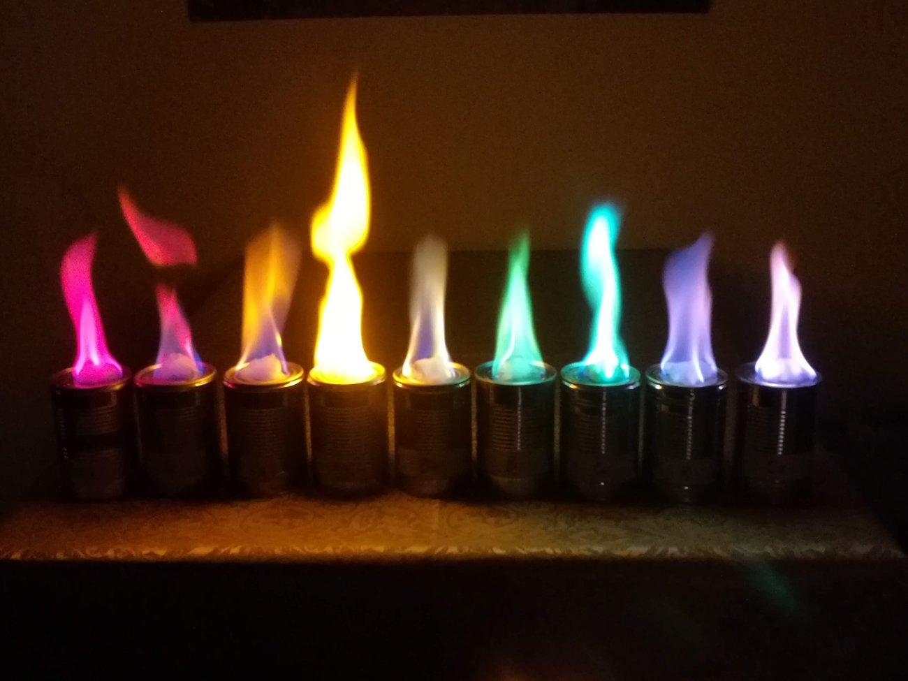 Próby płomieniowe: każdy metal alkaliczny barwi płomień inaczej — lit na karmazynowo, sód na żółto, potas na fioletowo. To spektralne „odciski palców" pierwiastków (na zdjęciu także Sr, Ca, Ba, Cu). · źródło: Hegelrast, Wikimedia Commons, CC BY-SA 4.0