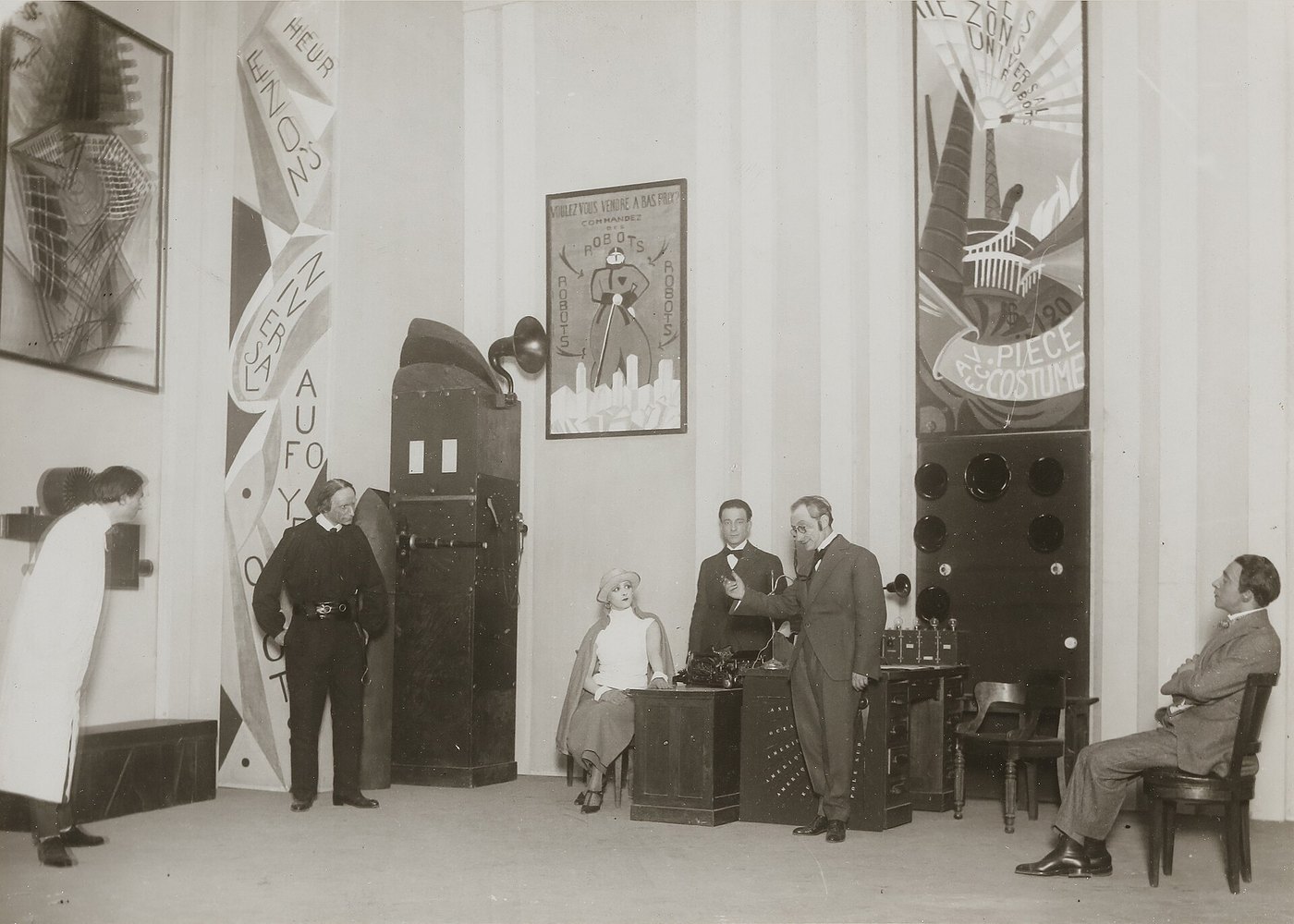 Scena z R.U.R. w Comédie des Champs-Élysées (Paryż, 26 marca 1924) — biuro fabryki, a na ścianie plakat reklamowy „Voulez-vous vendre à bas prix? Commandez des robots” · fot. Henri Manuel, źródło: Wikimedia Commons, domena publiczna